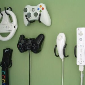 Könnte beinhalten: Eine Sammlung von Videospiel-Controllern und Zubehör, darunter ein weißes Lenkrad, ein weißer Xbox-Controller, ein schwarzer PlayStation-Controller, eine weiße Wii-Fernbedienung und ein schwarzer Gitarren-Controller, an einer grünen Wand.