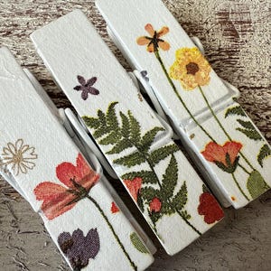 Puede incluir: Tres pinzas de ropa de madera blanca con diseños florales. Cada pinza presenta una flor diferente, incluyendo amapolas rojas, margaritas amarillas y helechos verdes. Las pinzas están dispuestas sobre una superficie texturizada de color marrón claro.