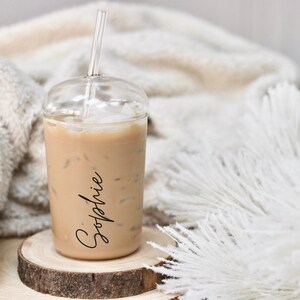 May include: A clear plastic iced coffee cup with a clear straw, filled with a light brown beverage and ice cubes. The name "Sophie" is written in black script on the side. The cup sits on a wooden coaster.