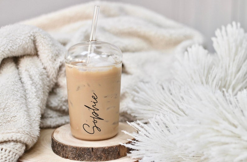 May include: A clear plastic iced coffee cup with a clear straw, filled with a light brown beverage and ice cubes. The name "Sophie" is written in black script on the side. The cup sits on a wooden coaster.