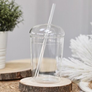 May include: A clear glass tumbler with a domed lid and a clear straw. The glass sits on a wooden coaster. A small potted plant and decorative white feathers are in the background.