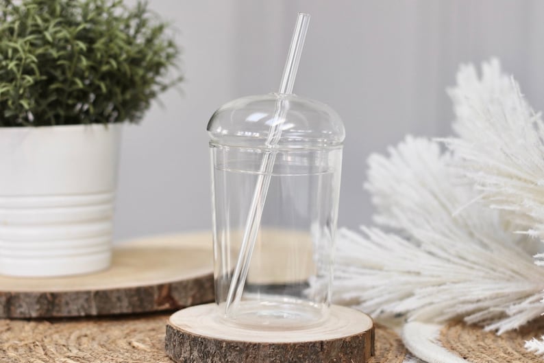 May include: A clear glass tumbler with a domed lid and a clear straw. The glass sits on a wooden coaster. A small potted plant and decorative white feathers are in the background.