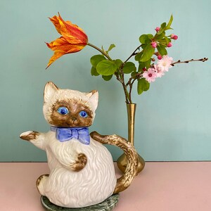 Peut inclure: Une théière en céramique en forme de chat siamois avec un corps crème, un visage et une queue bruns, des yeux bleu vif et un nœud papillon bleu. La théière repose sur une base circulaire en marbre vert à côté d'un vase en laiton fin.