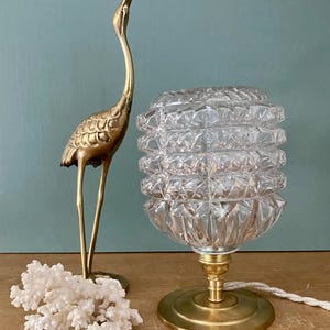 May include: A brass table lamp with a clear, faceted glass shade. The lamp has a round base and a cord.