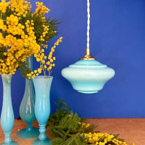 Peut inclure: Une lampe suspendue en verre bleu clair avec une base dorée est suspendue à un cordon blanc. Trois vases bleu clair contiennent des fleurs jaunes. Des fleurs jaunes et du feuillage vert sont disposés sur une surface en bois sur fond bleu.
