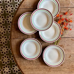 May include: A set of five white ceramic plates, each featuring a series of concentric rings in shades of deep red, bright green, and blue along the rim. The plates are arranged in a cluster, accompanied by a sprig of orange crocosmia flowers.