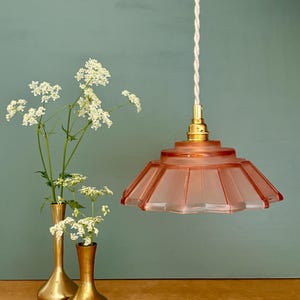 May include: A pink glass pendant light with a fluted shade hangs from a white twisted cord. The light fixture has a brass fitting. Two gold vases hold white flowers, set against a teal background.