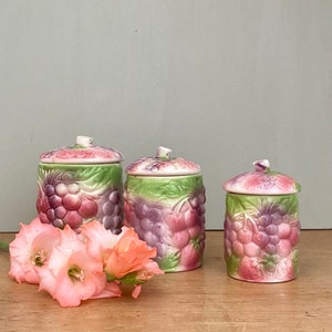 Könnte beinhalten: Set aus drei vintage Keramikbehältern mit Trauben- und Blattdesign. Die Behälter sind in den Farben Rosa, Grün und Lila.