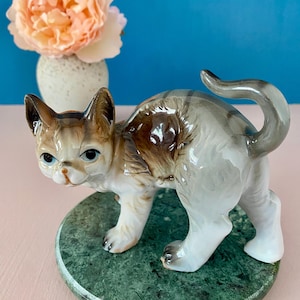 Może przedstawiać: Ceramiczna figurka kota z brązowymi, szarymi i białymi znaczeniami, przedstawiona w pozycji stojącej z wygiętym grzbietem i podniesionym ogonem. Kot jest umieszczony na okrągłej, polerowanej podstawie z zielonego marmuru o nakrapianej fakturze.