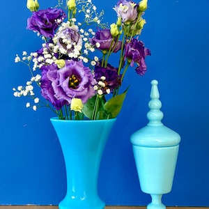 May include: A bright turquoise glass vase filled with purple eustoma flowers and white baby's breath, positioned next to a matching light blue glass apothecary jar with a tall, pointed lid and a pedestal base, both set against a solid blue backdrop.