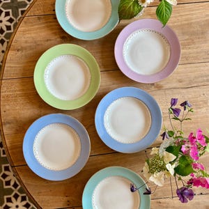 Puede incluir: Seis platos de cerámica en colores pastel con bordes dorados y delicados bordes interiores punteados están dispuestos sobre una mesa de madera. Los platos presentan bordes azul claro, verde, lavanda y turquesa. Hay flores en el fondo.