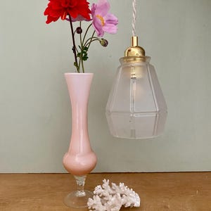 Könnte beinhalten: Eine gefrostete Glaspendelleuchte mit einer goldenen Metallfassung hängt an einem weißen Kabel. Eine rosa Glasvase mit roten und rosa Blumen steht auf einer Holzoberfläche neben einem weißen Korallenstück.
