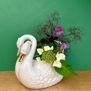 Pode incluir: Um vaso de cerâmica branca em forma de cisne com acabamento brilhante, detalhes de penas moldadas e detalhes dourados no bico. O cisne contém um arranjo floral composto por botões de hortênsia verdes, clematis roxas e folhagem escura.