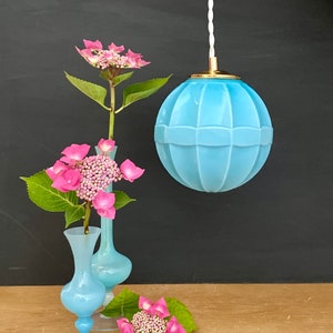 May include: A light blue, globe-shaped pendant light with a gold-colored cap hangs from a white cord. Two small, light blue vases hold pink hydrangea flowers. A few hydrangea blooms rest on a wooden surface, with a dark background.