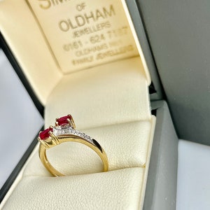 May include: A gold ring featuring two oval-cut red gemstones and small white gemstones, presented in a grey jewellery box. The box displays the text "SIMPSONS of OLDHAM JEWELLERS".