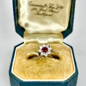 May include: A gold ring featuring a central red gemstone encircled by small white stones, in a floral arrangement. The ring is presented in a vintage blue jewellery box with a cream interior.