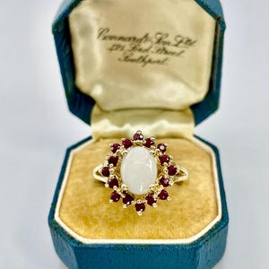 May include: A gold ring featuring an oval, off-white central stone, encircled by small, round, red gemstones. The ring is presented in a vintage blue jewellery box with a gold interior.