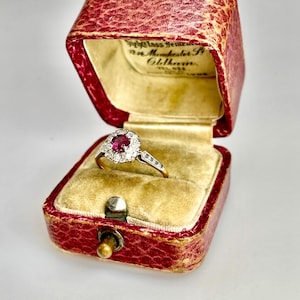 May include: A vintage ruby and diamond ring displayed in a red leatherette jewellery box. The ring has a central ruby surrounded by a halo of diamonds, set in a gold band. The box is lined with beige velvet.