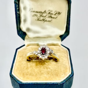 May include: A gold ring featuring a central oval-cut red ruby surrounded by a halo of small round diamonds, presented in a blue octagonal jewellery box with the text Connards Son Ltd 491 Lord Street Southport printed on the inner white satin lid.