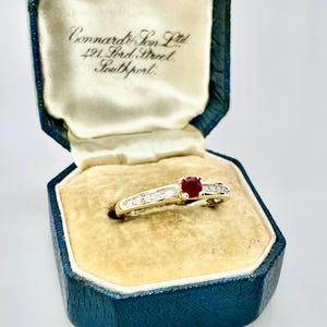 May include: A gold ring featuring a round red ruby centre stone flanked by channel-set diamonds on the band, presented in a dark blue velvet jewellery box with the text Connard & Son Ltd, 421 Lord Street, Southport printed on the inner lid.