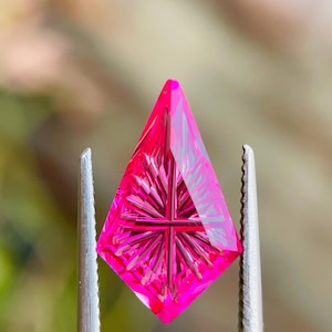 May include: A faceted, hot pink gemstone held by tweezers. The gem has a geometric shape with a cross-like design carved into its center. The background is blurred, highlighting the gem's vibrant color and intricate details.