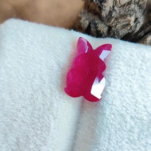 Könnte beinhalten: Ein facettierter, rubinroter Hase aus Edelstein. Der Edelstein liegt auf einer weißen, strukturierten Oberfläche. Der Hase hat spitze Ohren und einen abgerundeten Körper. Die Facetten des Edelsteins reflektieren das Licht.