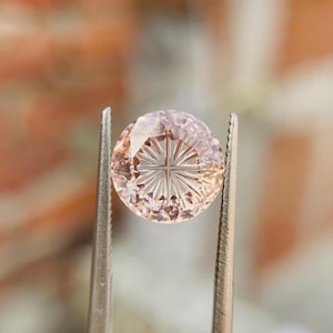 Puede incluir: Una piedra preciosa redonda de color melocotón pálido con un patrón de rayos de sol tallado con precisión en su superficie. La piedra está sujeta por dos pinzas metálicas, mostrando sus bordes facetados y las líneas radiantes grabadas.