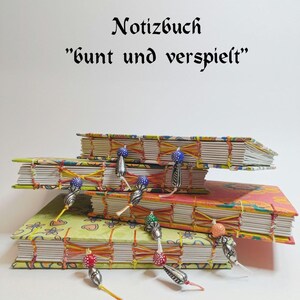 Könnte beinhalten: Drei farbenfrohe Notizbücher mit dekorativen Nähten und bunten Perlen. Die Notizbücher sind übereinander gestapelt. Das oberste Notizbuch trägt den Text "Notizbuch "bunt und verspielt"" in schwarzer Schrift.