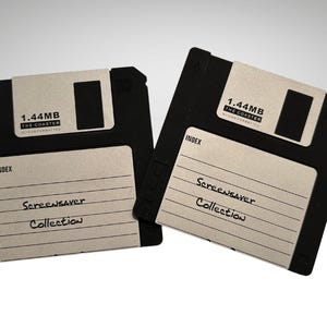 Pode incluir: Duas bases para copos pretas em forma de disquete com etiquetas brancas. As etiquetas dizem "1.44MB" e "Screensaver Collection". As bases para copos são projetadas para se assemelhar a dispositivos de armazenamento de computador clássicos.