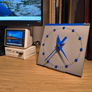 May include: A square, silver clock with blue hands and blue square markers. The clock is on a wooden surface, with a small computer monitor and a retro-style computer nearby. The clock's design is modern and minimalist.