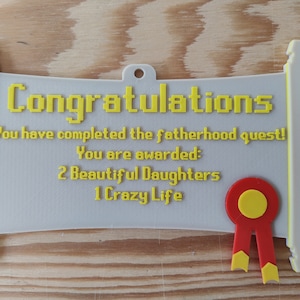 May include: A light grey scroll-shaped plaque with yellow text that reads: Congratulations, You have completed the fatherhood quest! You are awarded: 2 Beautiful Daughters, 1 Crazy Life. A red and yellow ribbon seal is attached to the bottom right.