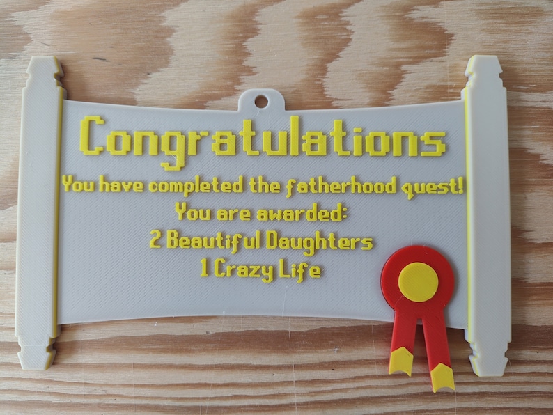 May include: A light grey scroll-shaped plaque with yellow text that reads: Congratulations, You have completed the fatherhood quest! You are awarded: 2 Beautiful Daughters, 1 Crazy Life. A red and yellow ribbon seal is attached to the bottom right.