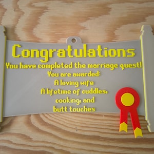 May include: A light grey scroll-shaped plaque with yellow pixelated text that reads: Congratulations, You have completed the marriage quest! You are awarded: A loving wife, A lifetime of cuddles, cooking, and butt touches. A red and yellow ribbon is attached.