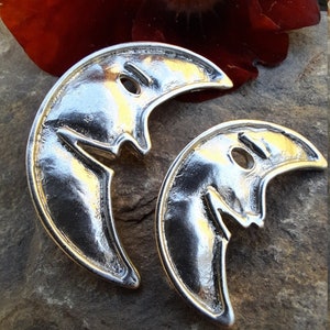 May include: Two silver crescent moon charms with a face-like design. The charms are made of metal and have a shiny finish.