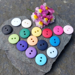 May include: Assortment of colourful plastic buttons in various shades of blue, green, pink, yellow, orange, purple, and white. The buttons are arranged in a heart shape on a grey stone surface.