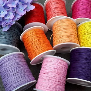 May include: Assortment of spools of colourful waxed cotton cord, in shades of purple, pink, orange, yellow, red, and grey. The spools are arranged on a textured surface.