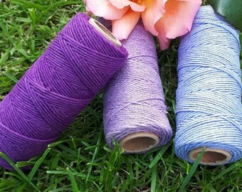 Peut inclure: Trois bobines de ficelle dans des tons de violet, lavande et bleu clair. Les bobines sont posées sur un lit d'herbe verte avec une rose rose en arrière-plan.