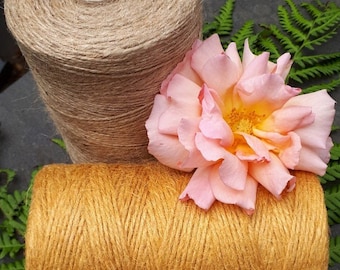 May include: Two spools of twine, one natural brown and one golden yellow, are displayed on a dark gray surface with green fern leaves. A pink rose is placed on top of the spools.