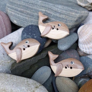 May include: Three wooden whale-shaped buttons with brown and tan coloring. The whales have small, round eyes and two holes for sewing. The buttons are arranged on a bed of smooth, gray and brown stones.