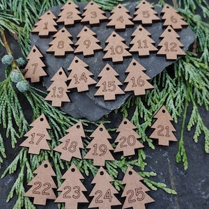 May include: Twenty-five wooden Christmas tree decorations, each numbered 1 to 25. The light brown decorations are arranged on a dark grey surface with green foliage.