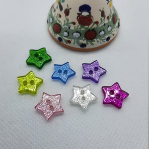 Può includere: Una collezione di bottoni a forma di stella in vari colori, tra cui verde, giallo, blu, viola, rosa e argento. Ogni bottone ha due fori per cucire e una finitura glitterata. Sullo sfondo una campana decorativa in ceramica.