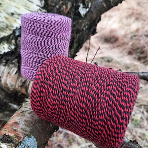 May include: Two spools of twine, one pink and black, the other red and black, with a wavy pattern. The spools are sitting on a tree branch.