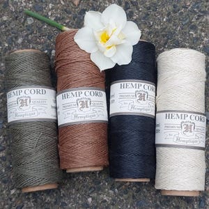 Könnte beinhalten: Vier Spulen Hanfschnur in Olivgrün, Braun, Schwarz und Creme, jeweils mit der Aufschrift "Hemp Cord" und "Hemptique". Eine weiße Narzisse mit gelbem Zentrum liegt obenauf. Die Spulen sind auf einer strukturierten Oberfläche angeordnet.