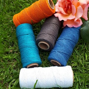 Peut inclure: Cinq bobines de ficelle de différentes couleurs : orange, bleu sarcelle, bleu foncé, gris et blanc. Les bobines sont disposées en cercle sur un lit d’herbe verte.