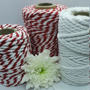 May include: Three spools of cotton twine in red and white, and solid white. The twine is tightly wound around cardboard cores. A white chrysanthemum flower is in front of the spools.