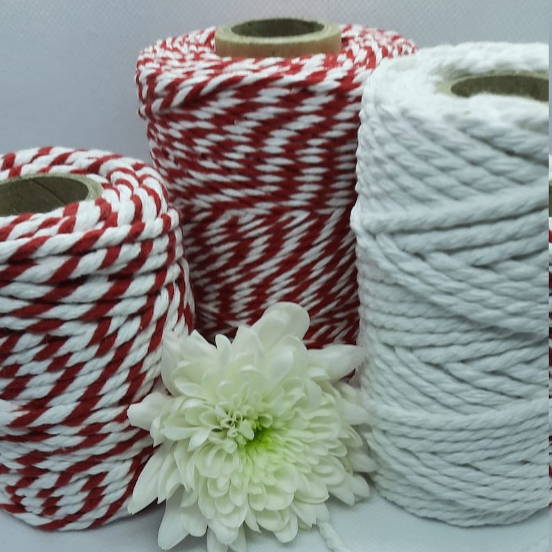 May include: Three spools of cotton twine in red and white, and solid white. The twine is tightly wound around cardboard cores. A white chrysanthemum flower is in front of the spools.