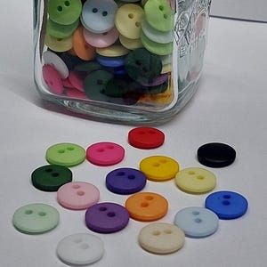 Könnte beinhalten: Ein durchsichtiges Glasgefäß gefüllt mit einer Vielzahl kleiner, runder Knöpfe mit zwei Löchern in verschiedenen Farben wie Rot, Blau, Grün, Gelb, Rosa, Lila, Orange, Schwarz und Weiß, mit einigen losen Knöpfen vor dem Glasgefäß.