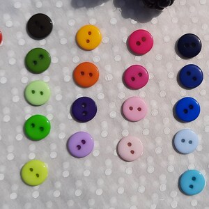 May include: Assortment of two-hole buttons in various colors, including red, green, blue, purple, pink, orange, yellow, and white. The buttons are arranged in a grid pattern on a white background.