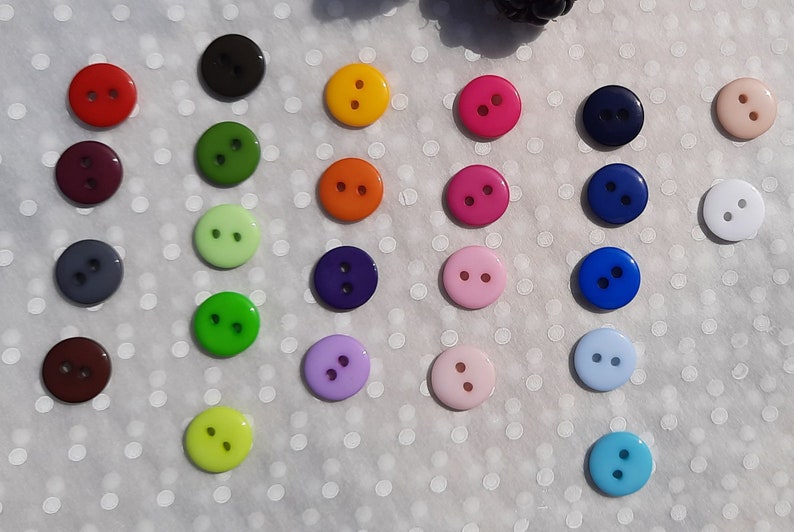 May include: Assortment of two-hole buttons in various colors, including red, green, blue, purple, pink, orange, yellow, and white. The buttons are arranged in a grid pattern on a white background.
