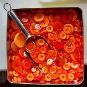 May include: A metal scoop sits in a rectangular tin overflowing with orange buttons. The buttons are in various shades of orange and red-orange. Some have four holes, others two, and some are decorative. The tin is filled with buttons.